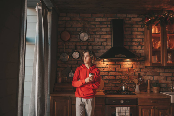 Sabahları uzun saçlı, evde mutfak hazırlayan genç bir çocuk. Havalı zoomer gen Z gündelik kıyafetlerle yeni yılı kutluyor Noel çelenkleri, yemek pişirmekten zevk alan xmas arifesi - Fotoğraf, Görsel
