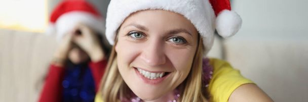 Kameraya gülümseyen, Noel Baba şapkası takan, Noel Baba 'nın yeni yıl partisinde arkadaşlarıyla buluşan çekici genç bir kadının portresi. Yeni yıl, tatil, eğlence konsepti. Bulanık arkaplan - Fotoğraf, Görsel