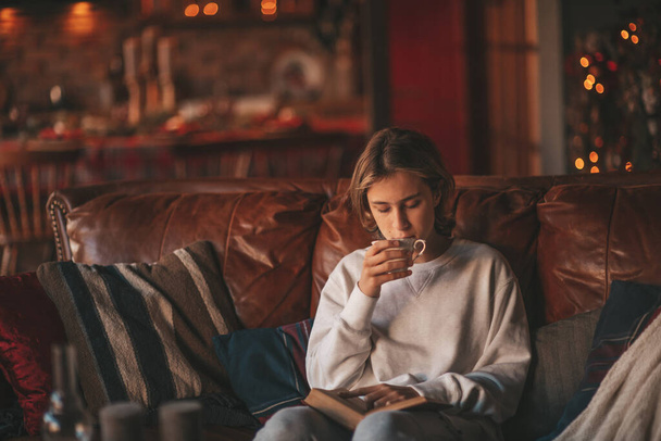 Young teen boy with long hair reading study book and enjoy hot drink distance learning at home. Stylish zoomer gen Z celebrates new year holidays with xmas tree bokeh lights garlands eve 25 december - Foto, Imagen
