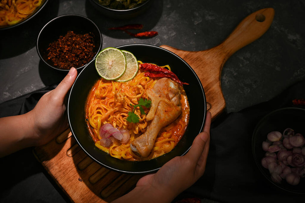 Manos de una hembra sosteniendo o sirviendo un tazón de Khao Soi Kai sobre la mesa. Curry amarillo cremoso y ligeramente picante con muslo de pollo. Concepto tradicional de cocina tailandesa - Foto, Imagen