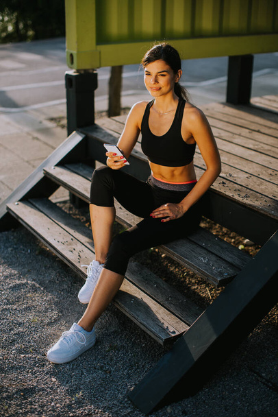 Vista a giovane donna che si prende una pausa durante l'esercizio al di fuori e utilizzando il telefono cellulare - Foto, immagini