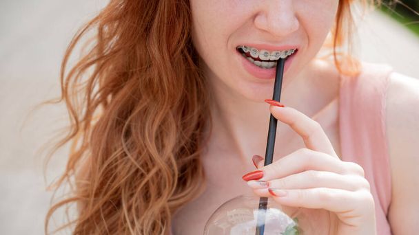 Young beautiful red-haired woman smiling with braces drinks cooling lemonade outdoors in summer. Cropped portrait Girl with freckles. - Foto, Imagen