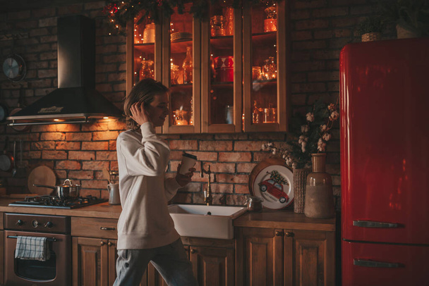 Young teenage boy with long hair in morning routine and preparing on kitchen at home. Stylish zoomer gen Z in casual clothing celebrates new year lights holidays garlands eve xmas enjoying cooking - Fotografie, Obrázek