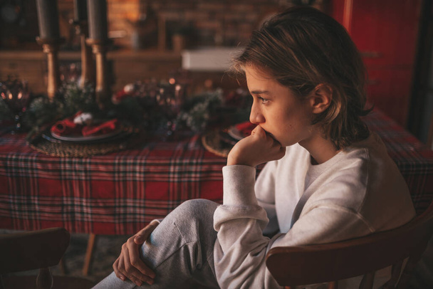 Young teen boy with long hair thoughtful look sad eyes negative mood angry and crying at home. Stylish zoomer gen Z pensive on new year holidays with xmas tree bokeh lights garlands eve 25 december - Foto, Imagen