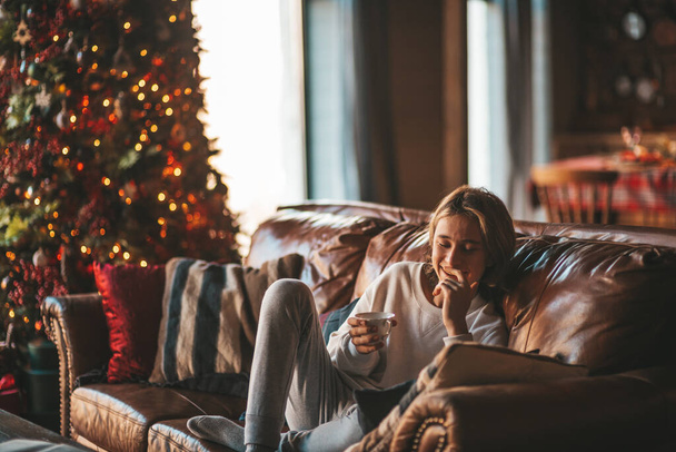 Young emotional cheerful teen boy with long hair laughter and joyful at cozy home. Stylish zoomer gen Z in good mood celebrates new year holidays with xmas tree bokeh lights garlands eve 25 december - Foto, afbeelding