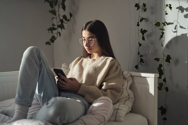Caucasian teenage girl browsing phone while sitting on bed - Zdjęcie, obraz