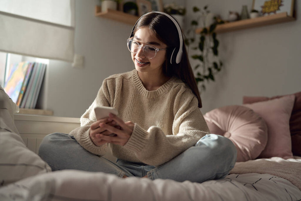Καυκάσιος έφηβος κορίτσι περιήγηση τηλέφωνο με χαμόγελο και φορώντας ακουστικά, ενώ κάθεται στο κρεβάτι - Φωτογραφία, εικόνα