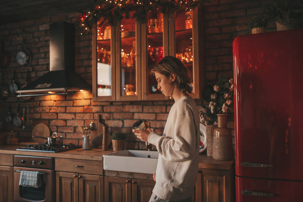 Молодой подросток с длинными волосами в утренней рутине и готовится на кухне дома. Стильный Zoomer ген Z в повседневной одежде празднует Новый год огни праздничные гирлянды накануне Рождества наслаждаясь приготовлением пищи - Фото, изображение