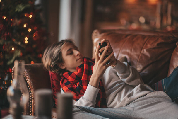 Young teen boy with long hair holding smartphone using wireless technology mobile device for chatting sms. Stylish child gen Z celebrates new year with xmas tree bokeh lights garlands eve 25 december - Photo, Image