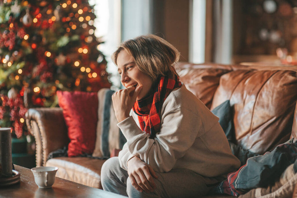 Young emotional cheerful teen boy with long hair laughter and joyful at cozy home. Stylish zoomer gen Z in good mood celebrates new year holidays with xmas tree bokeh lights garlands eve 25 december - Photo, Image