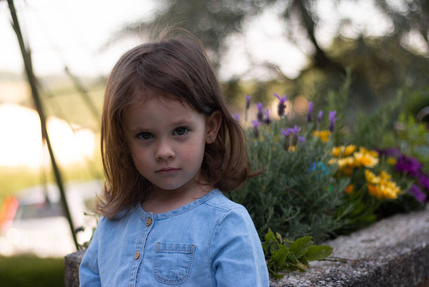 A little beautiful preschooler girl in a denim dress stands on a blurred background near bright flowers - Photo, Image
