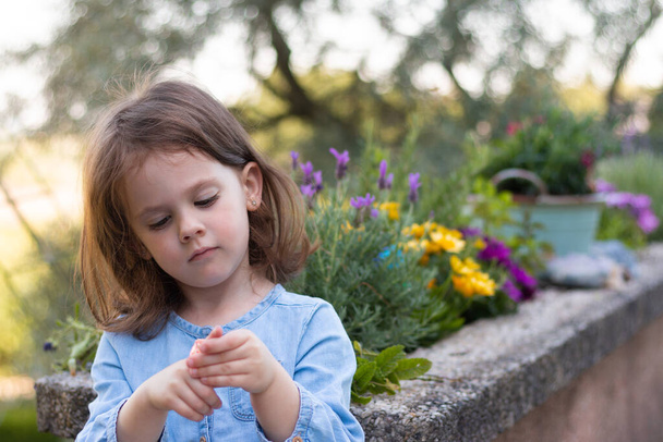 Pieni kaunis esikoululainen tyttö denim-mekossa seisoo hämärtyneellä taustalla lähellä kirkkaita kukkia. - Valokuva, kuva