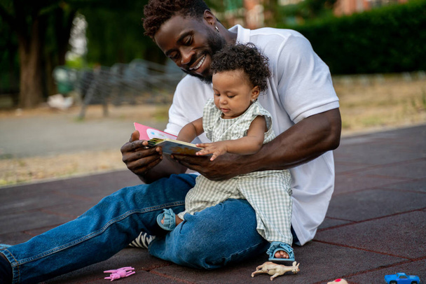 Child seated on the lap of her happy parent touching an open book in his hands - Photo, Image