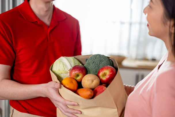 Asian delivery man in red uniform giving paper bag of food, fruit, vegetable to female customer to her house. Delivering grocery service at home - Foto, Imagen