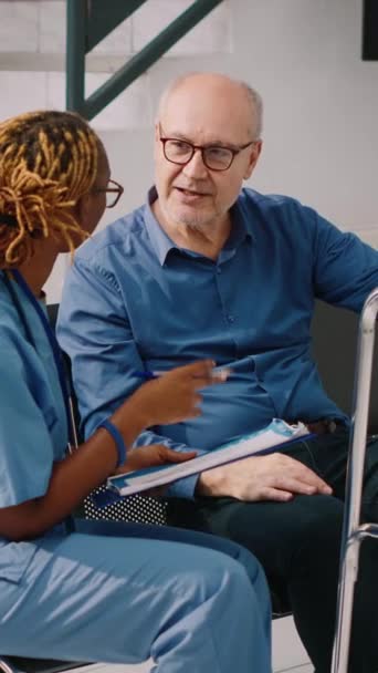Вертикальне відео: Медичний асистент консультує літнього чоловіка з прогулянковою рамкою, сидячи в холі прийому госпіталю. Медсестра розмовляє зі старою людиною в залі очікування в медичному центрі, заповнюючи клінічну
 - Кадри, відео