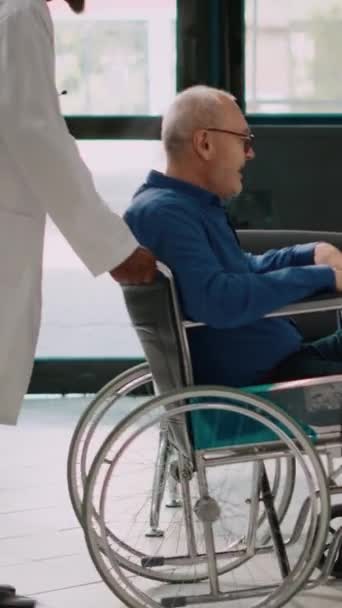 Pystysuora video: Lääkintämies tuo vanhempi mies pyörätuolissa puhua naisen ja lapsen, on terve ja alkaa kävellä kävelykehys. Iäkäs potilas toipumassa terveyskeskuksessa. - Materiaali, video