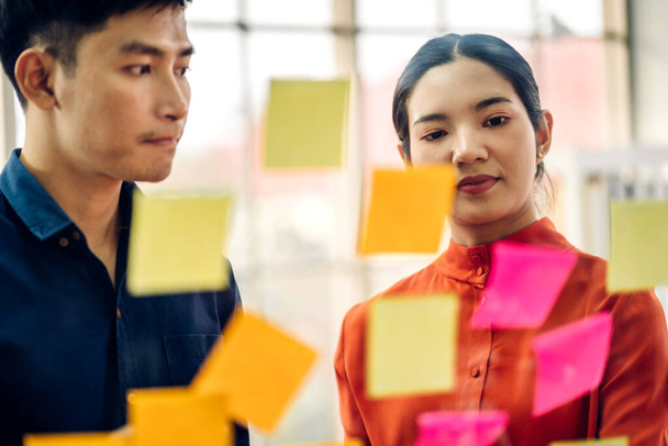 Casual creative happy two smile asian business people plan strategy analysis and brainstorm putting post it sticker note to share idea for startup project on glass board at modern office - Photo, Image