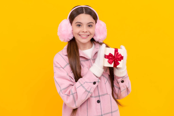 smiling child in earmuffs and mittens hold present box on yellow background, purchase. - Fotó, kép