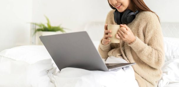Young smiling happy beautiful asian woman relaxing using laptop computer in the bedroom at home.Young creative girl working and typing on keyboard.work from home concept - Фото, изображение