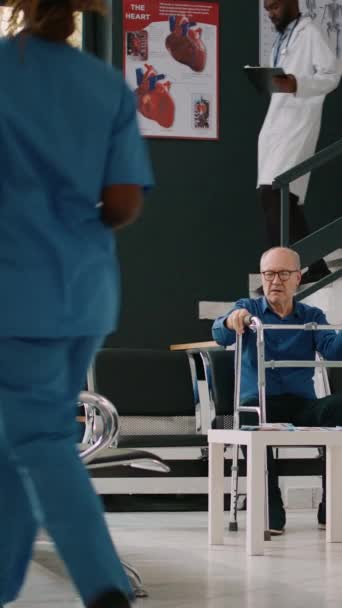Pystysuora video: Vanha potilas puhuu ihmisille odotushuoneen aulassa, käyttäen kävelykehystä terveyskeskuksen vastaanotossa. Vanhempi mies, jolla on vamma ja joka odottaa yleislääkärin kuulemista - Materiaali, video