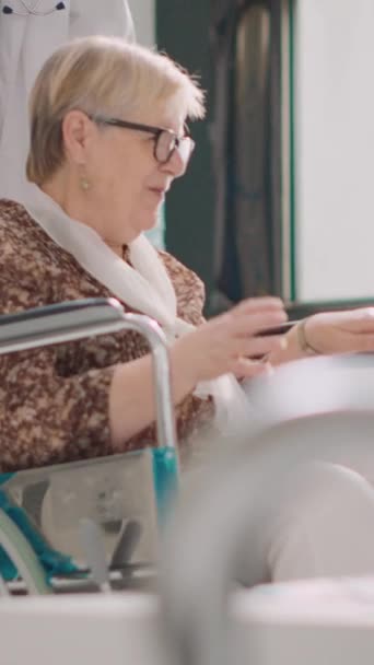 Függőleges videó: Tolószékben ülő idős személy járókerettel az egészségügyi központban, a kezelés után egészségesnek érzi magát. Férfi orvos hozza idős beteg az előcsarnokban, hogy találkozzon a kislány - Felvétel, videó