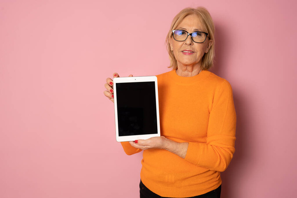 ピンクの背景に隔離されたタブレットコンピュータを使用して笑顔の美しい高齢者の成熟した女性の肖像画。テキストのコピースペース. - 写真・画像