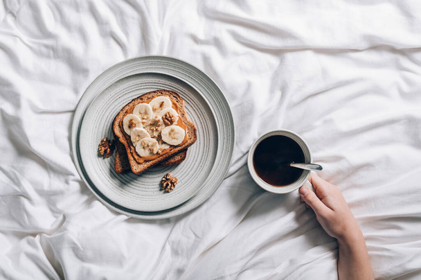 Fıstık ezmeli, muzlu ve ezilmiş cevizli kepekli ekmeğin açık sandviçi. Yatakta kahvaltı tabakta ve yatakta kahve. - Fotoğraf, Görsel