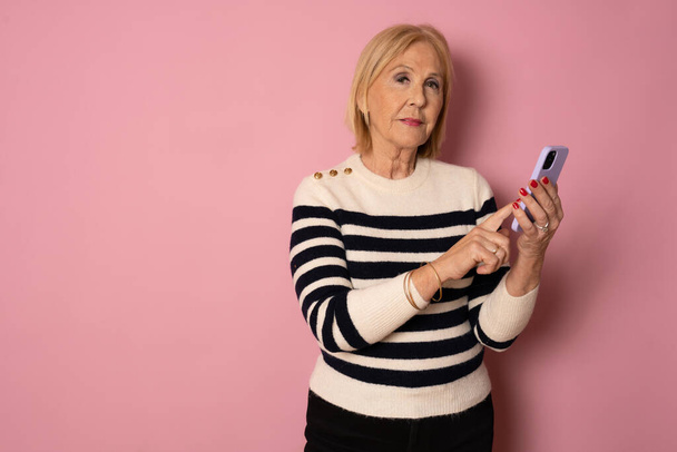 Feliz madura mujer mayor sosteniendo teléfono inteligente utilizando aplicaciones móviles en línea, sonriendo anciana señora sms mensajes de texto de chat en el teléfono mirando a la cámara aislada en el fondo rosa - Foto, imagen