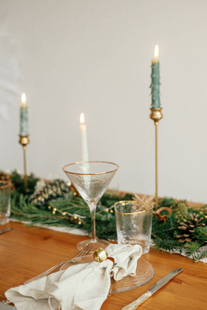 Christmas table setting. Linen napkin with bell on plate, vintage cutlery, glasses, fir branches with golden lights and pine cones on table. Holiday arrangement of table - Photo, image