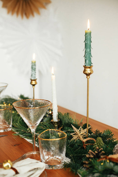 Stilvolle weihnachtliche Tischdekoration. Leinenserviette mit Glocke auf Teller, Vintage-Besteck, Weinglas, Tannenzweige mit goldenen Lichtern, Tannenzapfen und Kerze auf dem Tisch. Stimmungsvoller Feiertagsbrunch - Foto, Bild