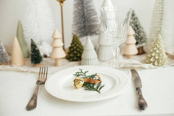 Stijlvolle kersttafel. Cedertak met belletje op bord, vintage bestek, wijnglas, moderne kerstbomen en huizen op witte rustieke tafel. Vakantie brunch, nieuwjaarsviering - Foto, afbeelding