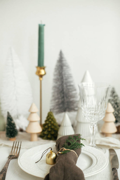 Stylish Christmas table setting. Cotton napkin with bell and fir on plate, vintage cutlery, wineglass, modern christmas trees and candle on white rustic table. Holiday brunch - Photo, Image