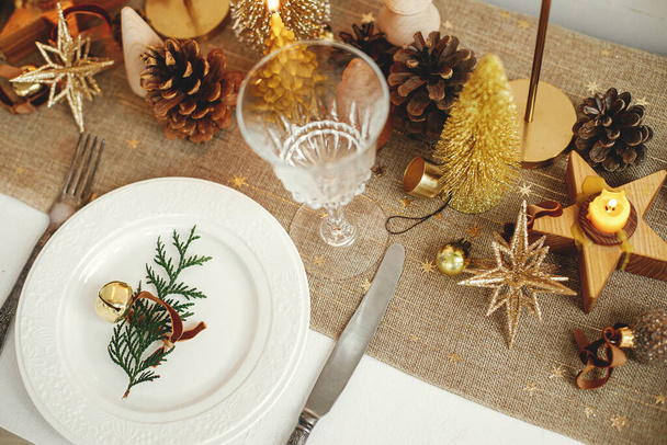 Kerst luxe tafel dekken. Cedertak met belletje op bord, vintage bestek, glazen, gouden kerstboompjes en ornamenten op tafel. Vakantie arrangement van tafel - Foto, afbeelding