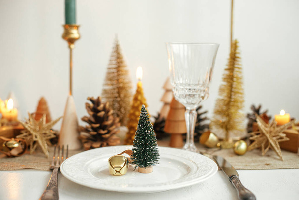 Christmas luxury table setting. Little Christmas tree with bell on plate, vintage cutlery, glasses, golden little christmas trees and ornaments on table. Holiday arrangement of table - Photo, Image