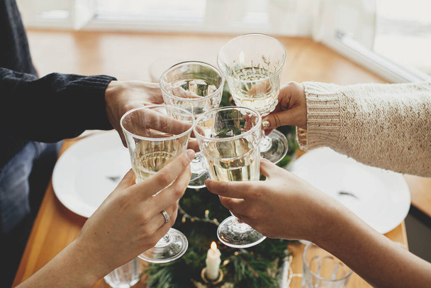 Χριστουγεννιάτικη γιορτή. Χέρια με ποτήρια κρασιού ψήσιμο και κάνοντας κλικ στο φόντο του καλαίσθητου τραπεζιού ρύθμιση με κλαδιά ελάτης με χρυσά φώτα και κεριά. Φίλοι γιορτάζουν με σαμπάνια - Φωτογραφία, εικόνα