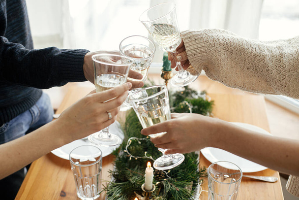Χέρια με ποτήρια σαμπάνιας κάνοντας κλικ στο φόντο του καλαίσθητου τραπεζιού με κλαδιά ελάτης με χρυσά φώτα και κεριά. Φίλοι κάνουν πρόποση με σαμπάνια και γιορτάζουν. Χριστουγεννιάτικη γιορτή - Φωτογραφία, εικόνα