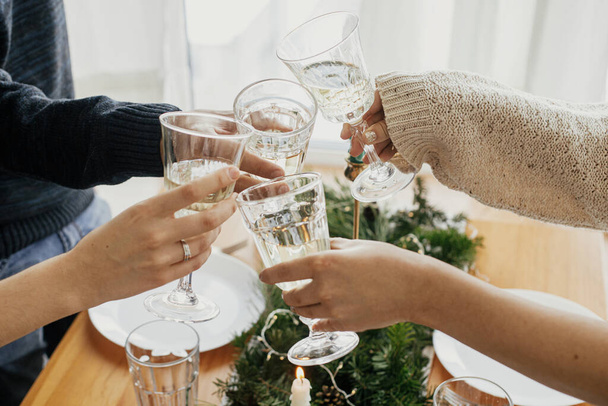 Christmas feast. Hands with wine glasses toasting and clicking on background of stylish table setting with fir branches with golden lights and candles. Friends celebrating with champagne - Zdjęcie, obraz