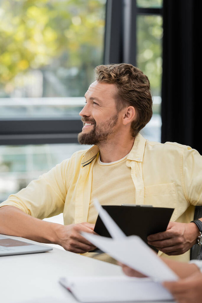 happy bearded businessman sitting with clipboard and looking away in office - Photo, Image