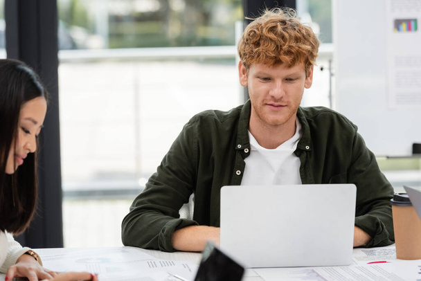 young redhead businessman working at laptop near asian colleague - Photo, image