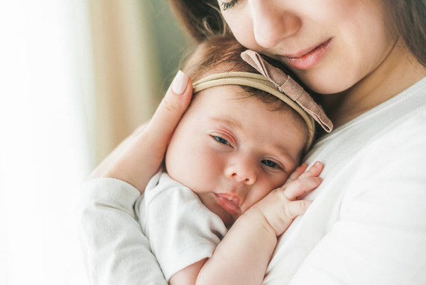 Szoros portré egy aranyos, sötét hajú újszülött lányról fehér öltönyben egy fiatal anya karjaiban. Anyaság. Szülő. Egészséges alvás. Otthon.. - Fotó, kép