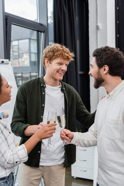 Smiling redhead businessman talking to interracial colleagues with champagne in office  - Fotoğraf, Görsel