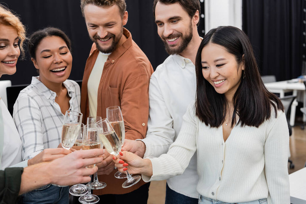 Smiling asian businesswoman clinking champagne with multiethnic colleagues in office  - Фото, изображение
