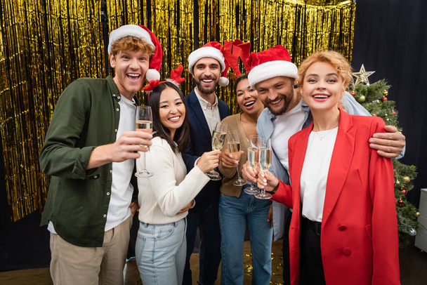 Emozionati uomini d'affari multietnici in cappelli di Babbo Natale con champagne vicino orpelli in carica  - Foto, immagini