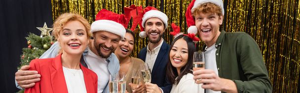 Cheerful interracial business people in santa hats holding champagne near tinsel in office, banner  - Foto, afbeelding