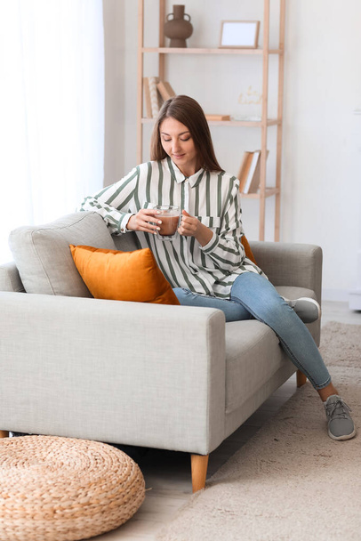 Giovane donna con tazza di caffè in vetro seduta su divano grigio a casa - Foto, immagini