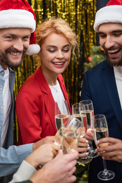 Businesswoman clinking champagne with smiling interracial colleagues in santa hats in office  - Photo, Image