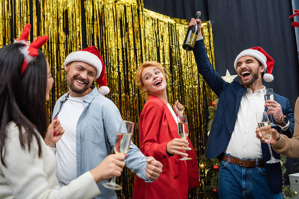 Excited businessman in santa hat holding bottle of champagne near interracial colleagues dancing in office  - 写真・画像