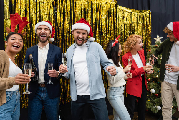 Весёлые многонациональные бизнесмены в шляпах Санта-Клауса держат шампанское рядом с коллегами и мишурой в офисе  - Фото, изображение