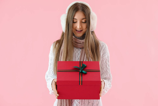 Happy young woman in ear muffs with Christmas gift on pink background - Photo, Image