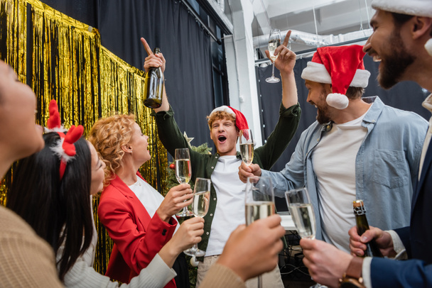 Aufgeregter Geschäftsmann mit Weihnachtsmütze hält Champagner in der Hand, während er Weihnachten mit multiethnischen Amtskollegen feiert  - Foto, Bild